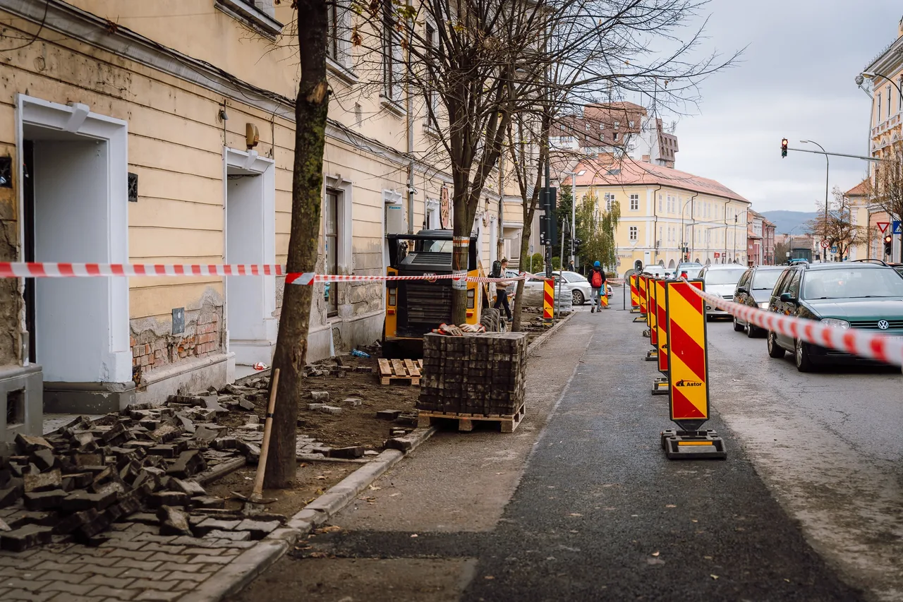 Utcai járdaépítési munkálatok láthatók egy régi épület mellett, felszedett kockakő halmokkal, egy kisebb munkagéppel, piros-fehér csíkos figyelmeztető szalaggal és sárga-piros forgalomelválasztó táblákkal az úton. A háttérben parkoló autók és sétáló emberek láthatók.