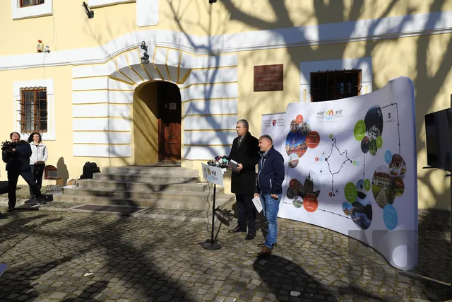 Sajtótájékoztató egy régi épület előtt a Vártemplom közelében: két férfi mikrofonok előtt beszél, mellettük „Visit Mures” feliratú turisztikai roll-up áll, Maros megye látványosságait bemutató térképpel és fotókkal. Baloldalt egy operatőr és egy nő figyeli az eseményt.