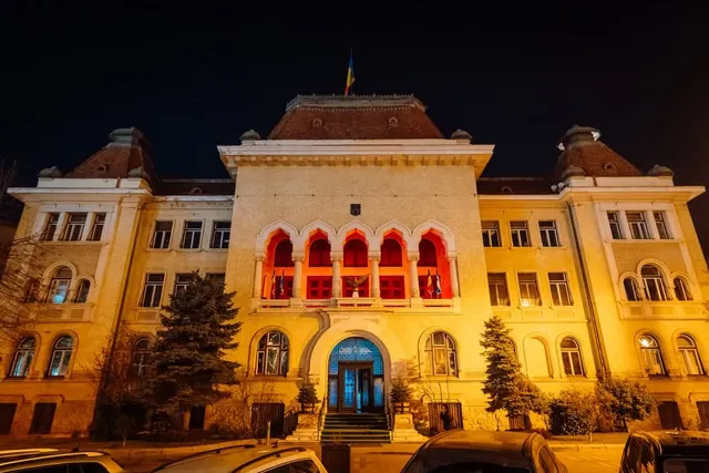 A marosvásárhelyi polgármesteri hivatal épülete éjszaka, homlokzati díszkivilágítással, a középső loggia és erkélyrészek piros fényben; az épület tetején román zászló, előtte fenyők és parkoló autók.