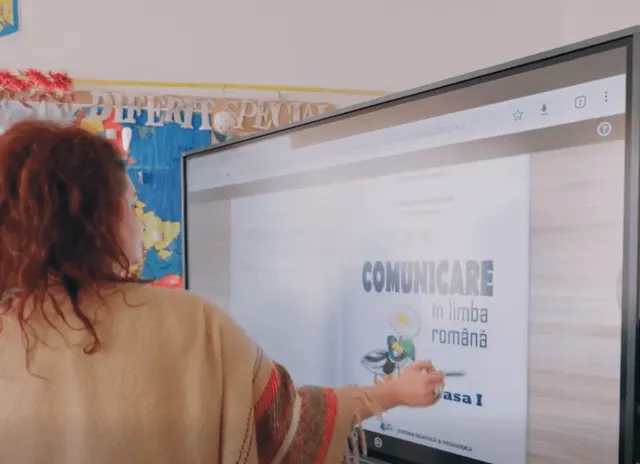 Egy pedagógus hátulról fotózva egy nagy interaktív kijelzőt kezel, amelyen egy „Comunicare în limba română” (Kommunikáció románul) című, első osztályos tankönyv digitális oldala látható.