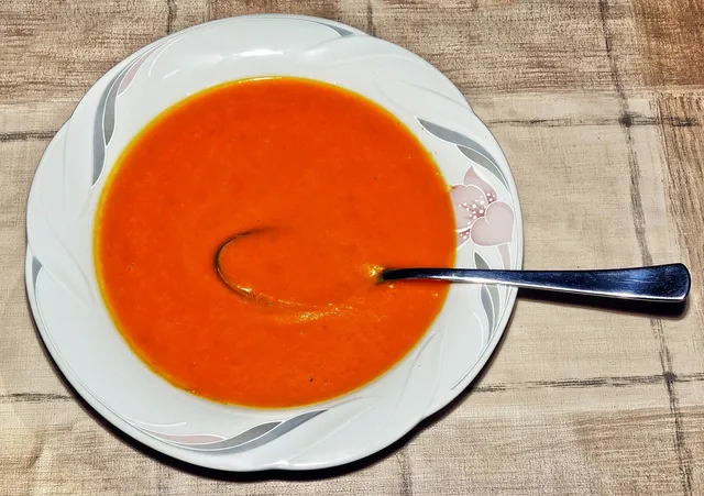 Fehér, virágmintás tányérban narancsvörös krémleves (feltehetően paradicsomleves), benne evőkanál, kockás mintás terítőn.