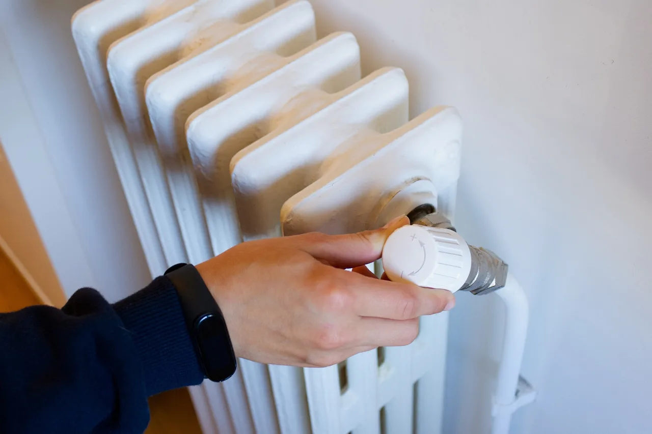 Egy kéz beállítja egy fehér öntöttvas fűtőtest termosztátszelepét, csuklóján fekete okoskarkötő látható.