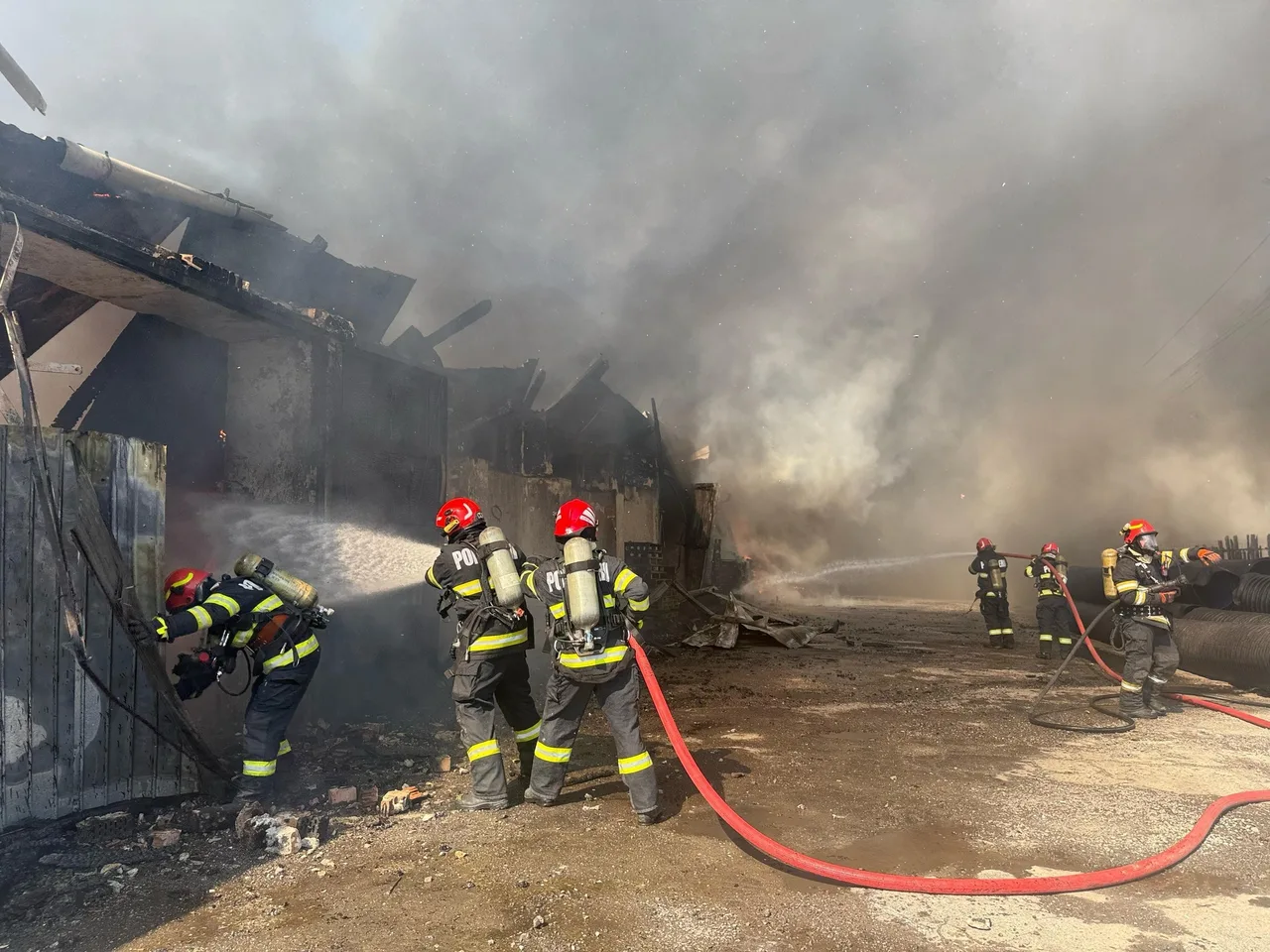 Több tűzoltó dolgozik egy leégett, füstölgő épület helyszínén, oltóvízzel próbálják eloltani a parazsat és megakadályozni a tűz továbbterjedését.