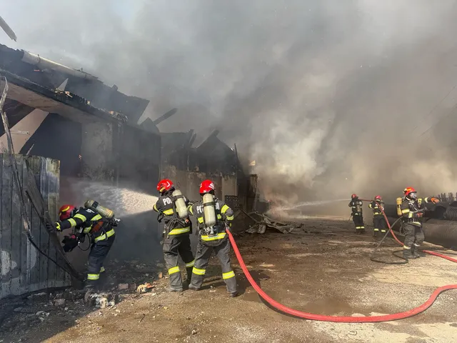 Több tűzoltó dolgozik egy leégett, füstölgő épület helyszínén, oltóvízzel próbálják eloltani a parazsat és megakadályozni a tűz továbbterjedését.