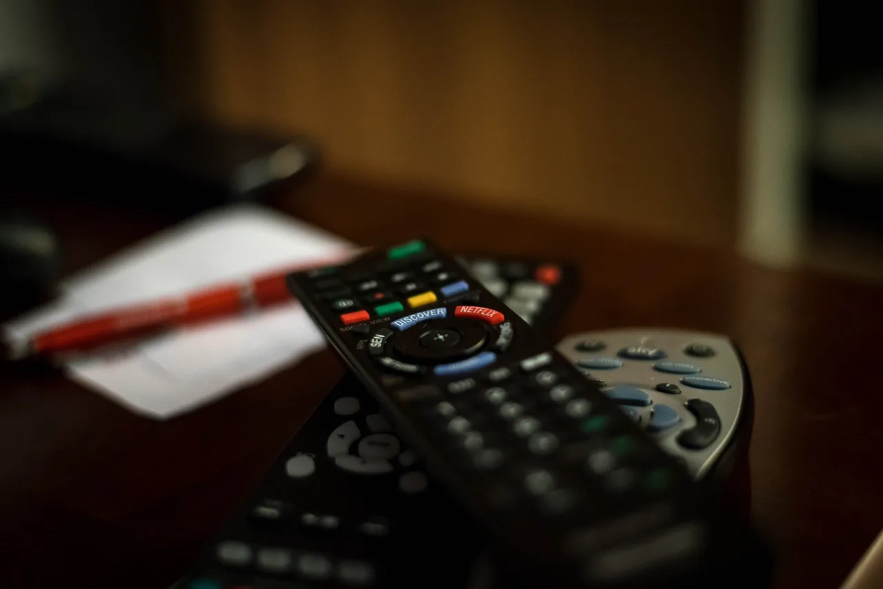 Több televíziós távirányító hever egymáson egy asztalon, előtérben egy Netflix és Discovery gombokkal ellátott fekete távirányító, háttérben elmosódva egy toll és egy papírlap látszik.