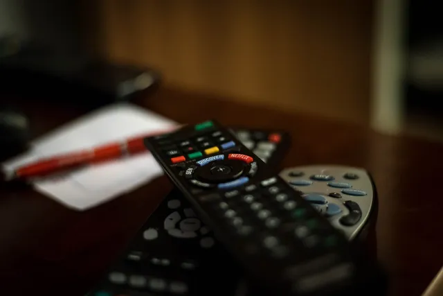 Több televíziós távirányító hever egymáson egy asztalon, előtérben egy Netflix és Discovery gombokkal ellátott fekete távirányító, háttérben elmosódva egy toll és egy papírlap látszik.