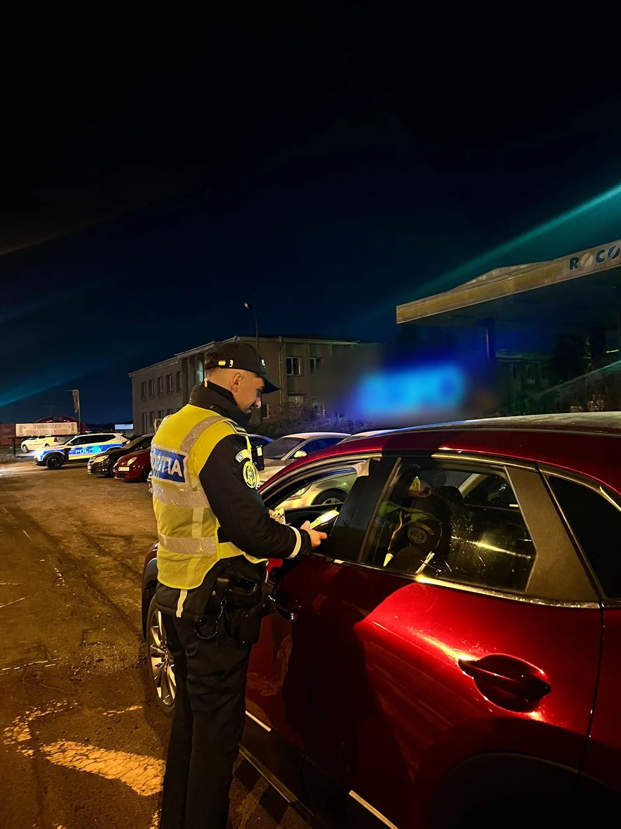 Éjszaka egy rendőr sárga láthatósági mellényben egy bordó autó vezetőjének ellenőrzi az okmányait egy parkolóban, háttérben rendőrségi autók és egy üzemanyagkút látszik.