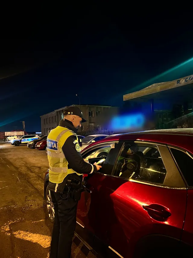 Éjszaka egy rendőr sárga láthatósági mellényben egy bordó autó vezetőjének ellenőrzi az okmányait egy parkolóban, háttérben rendőrségi autók és egy üzemanyagkút látszik.