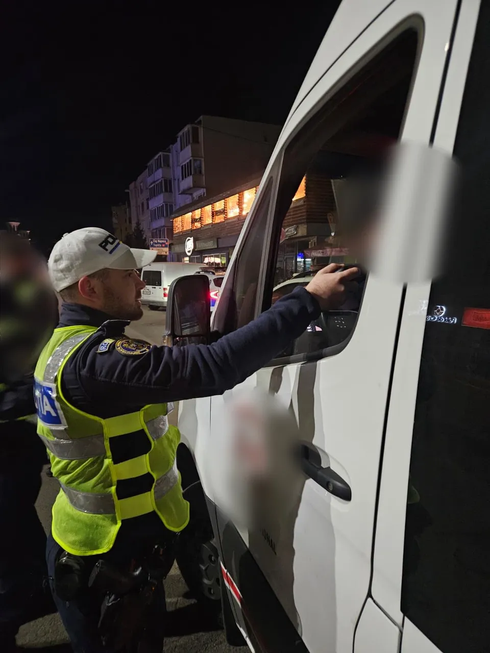 Éjjel egy sárga láthatósági mellényt és fehér sapkát viselő rendész nyújtogat egy fehér furgon vezetőülétébe, ahol egy elmosott alak ül; a háttérben lakótelepi épületek és boltok világítása látható. A kép egy közúti ellenőrzés pillanatképe.