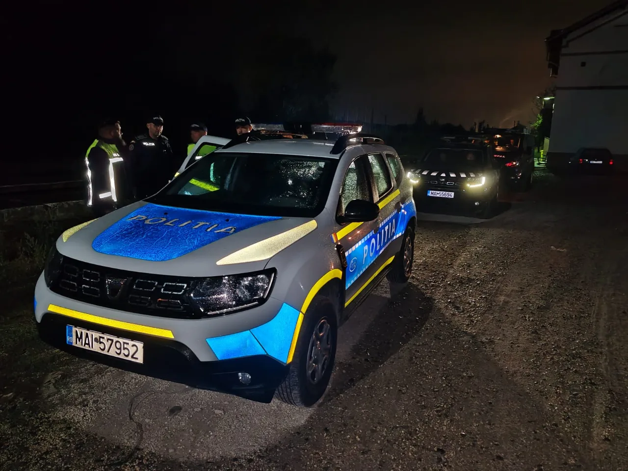 Éjszakai jelenet, előtérben egy rendőrségi terepjáró (Dacia Duster) „Politia” feliratokkal, háttérben további rendőrautók és egyenruhás rendőrök állnak egy csoportban egy vidéki úton, ház mellett.