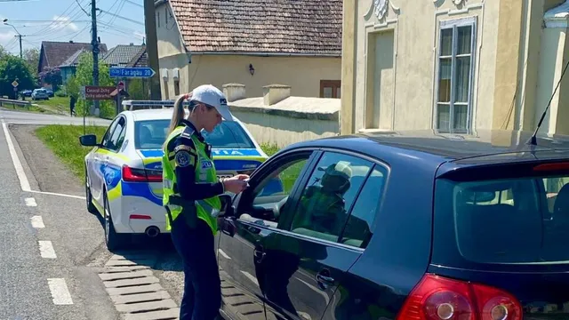 Rendőrnő láthatósági mellényben és sapkában igazoltat egy sötétkék autó sofőrjét egy falusi úton, mellette rendőrségi jelzőautó áll.