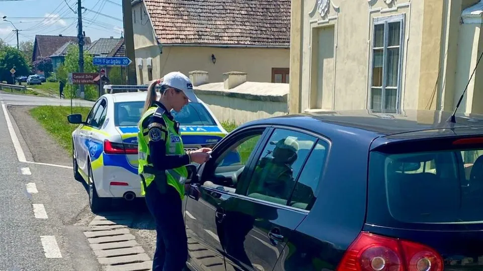Rendőrnő láthatósági mellényben és sapkában igazoltat egy sötétkék autó sofőrjét egy falusi úton, mellette rendőrségi jelzőautó áll.