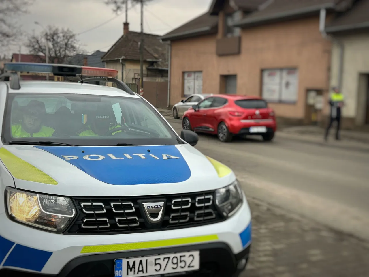 Rendőrségi Dacia Duster terepjáró áll egy utca szélén, „POLITIA” felirattal, benne két rendőr látható; a háttérben egy piros és egy szürke autó parkol az úton, valamint egy sárga-fekete láthatósági mellényt viselő rendőr áll az utcán.