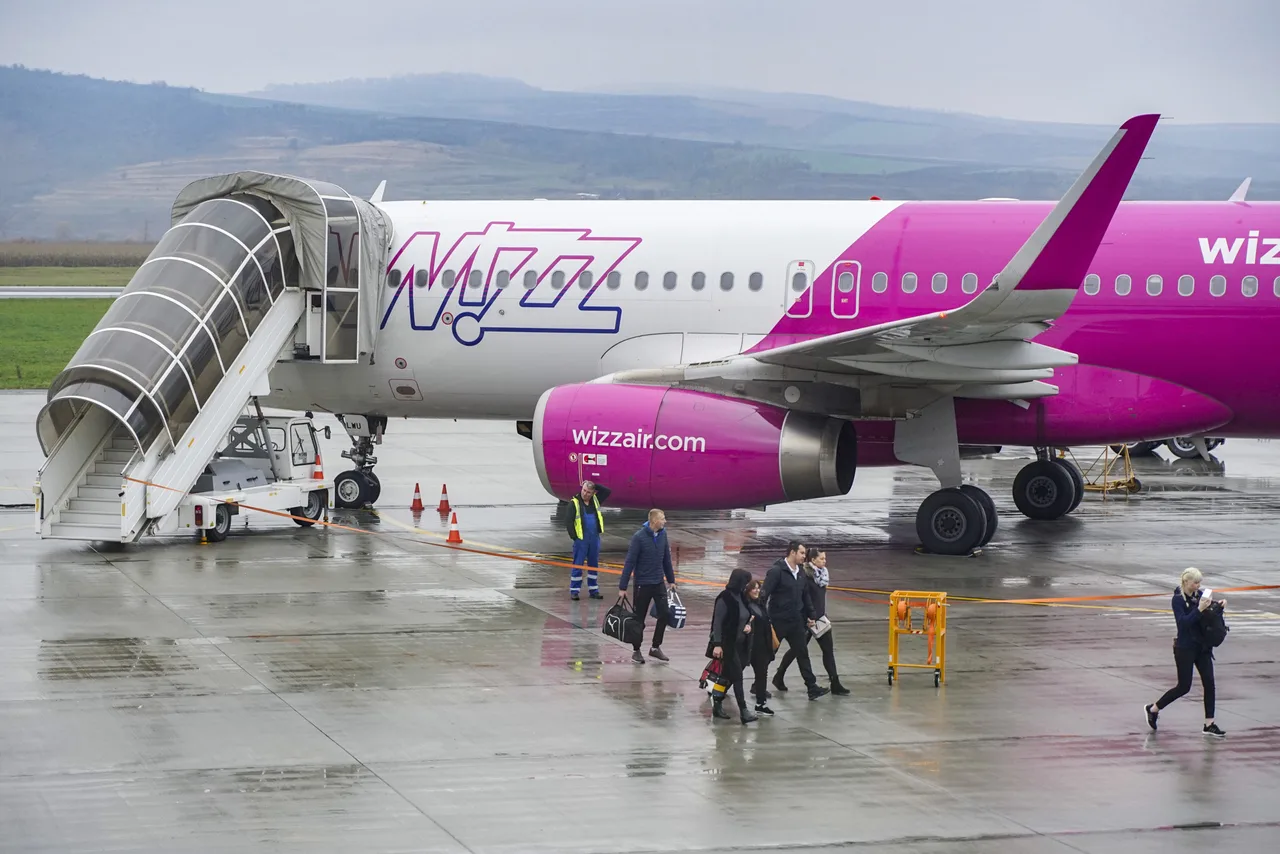 Wizz Air utasszállító repülőgép áll a repülőtér kifutópályáján, mellette kigördített lépcső, utasok szállnak le és sétálnak a leszállópályán csomagjaikkal esős időben.