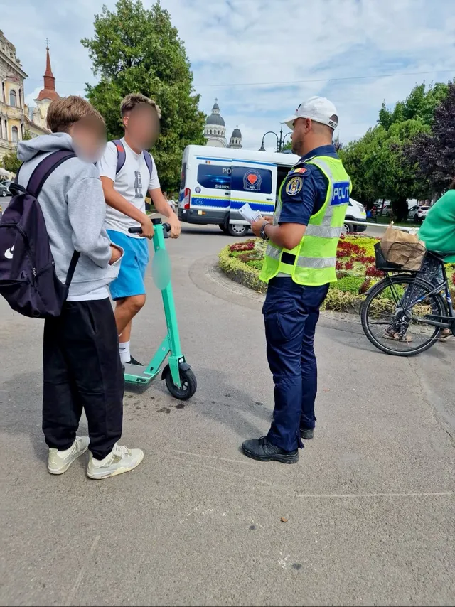Egy sárga-kék láthatósági mellényt és fehér sapkát viselő rendőr két, elektromos rollerrel közlekedő fiatalt szólít meg és ellenőriz egy közterületen. A háttérben rendőrségi kisbusz, virágágyás, kerékpáros és fák láthatók.