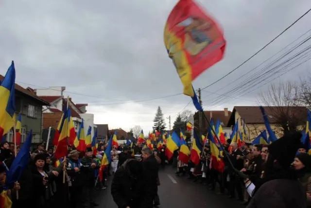 Utcán vonuló tömeg román zászlókkal és egy nagy piros-sárga-kék történelmi zászlóval (Moldva címerével), az emberek téli ruhában, egyesek népviseletben vonulnak egy falusias utcán.