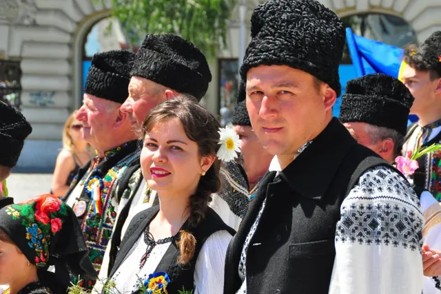 Hagyományos román népviseletbe öltözött férfiak és nők csoportja látható szabadtéri rendezvényen, a férfiak fekete kucsmában és mellényben, a nők hímzett fehér blúzban.
