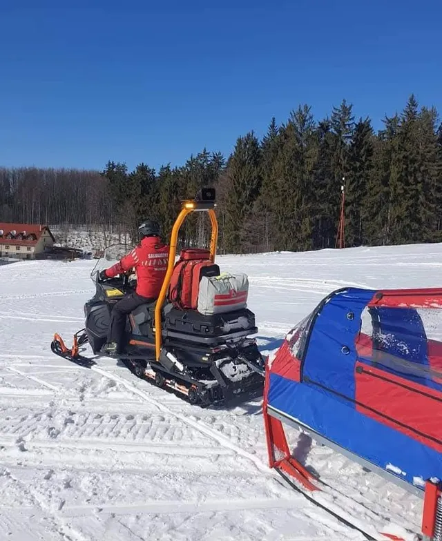 Piros dzsekit és sisakot viselő személy hómobilon (motoros szánon) húz egy lezárt, piros-kék színű mentőszánt havas terepen, háttérben fenyőerdő és tiszta kék ég.