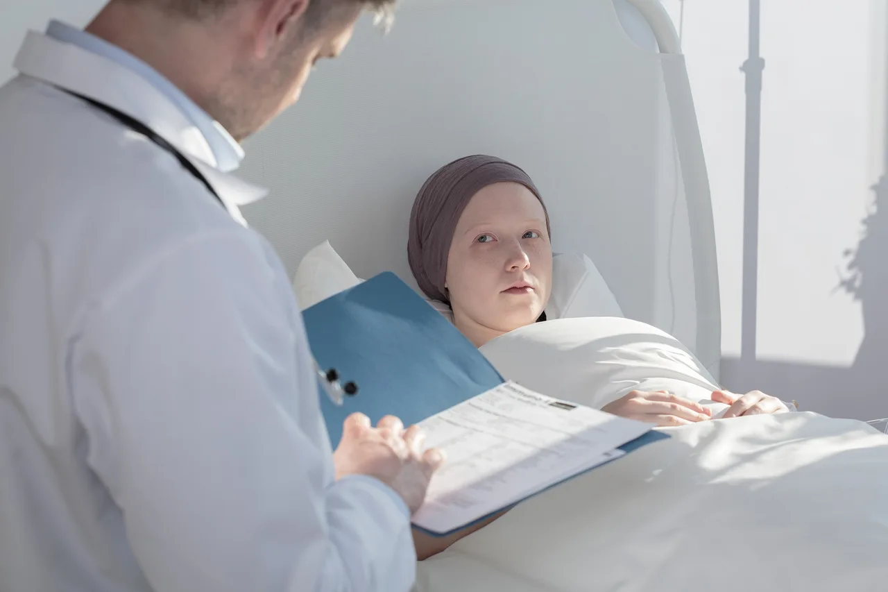 Kórházi ágyban fekvő, fejkendőt viselő fiatal beteg néz fel az orvosra, aki kék dossziéban lévő papírokat tanulmányoz mellette.