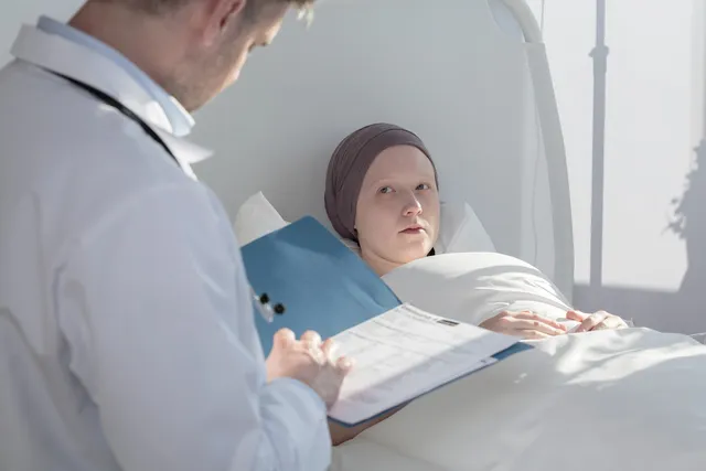 Kórházi ágyban fekvő, fejkendőt viselő fiatal beteg néz fel az orvosra, aki kék dossziéban lévő papírokat tanulmányoz mellette.