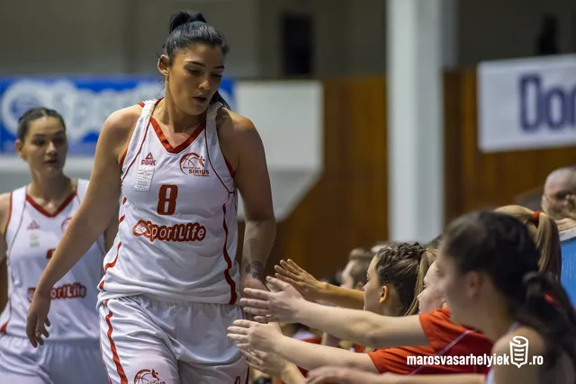 Kosárlabdázónő a Sirius Targu Mures fehér-piros mezében, 8-as számmal, sportcsarnokban vonul be a pályára, miközben a nézőtéren ülők ütik vele tenyerüket üdvözlésképp. Mögötte csapattársa is látszik hasonló mezben.