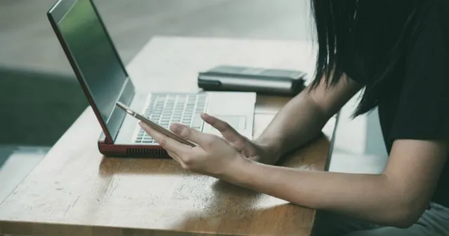 Egy asztalnál ülő személy laptop előtt okostelefont tart a kezében, mellette egy sötét pénztárca vagy tok látható az asztalon.