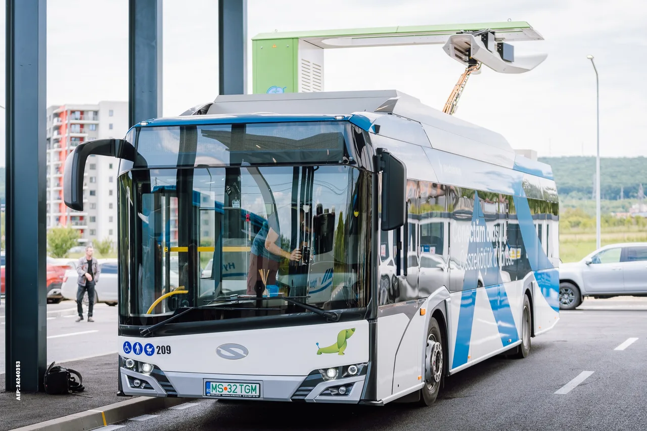 Elektromos töltőállomás felett álló, feltöltőkarral csatlakoztatott villanybusz egy megállóban, a sofőr a vezetőülésben látható, mellette a járdán egy férfi közelít a buszhoz.