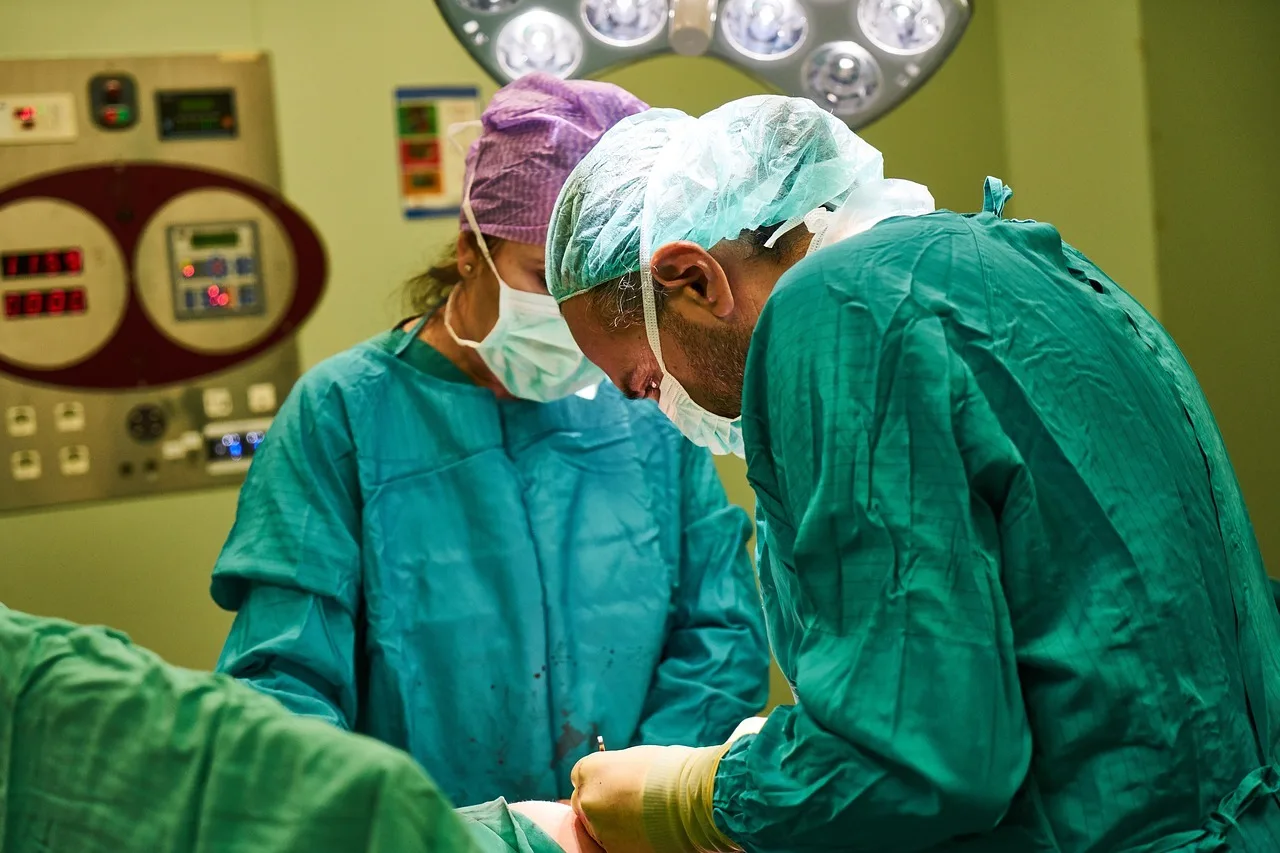 Két sebész műtői sapkában, maszkban és zöld műtői köpenyben hajol a beteg fölé egy műtőlámpa alatt, koncentráltan végzik a beavatkozást.