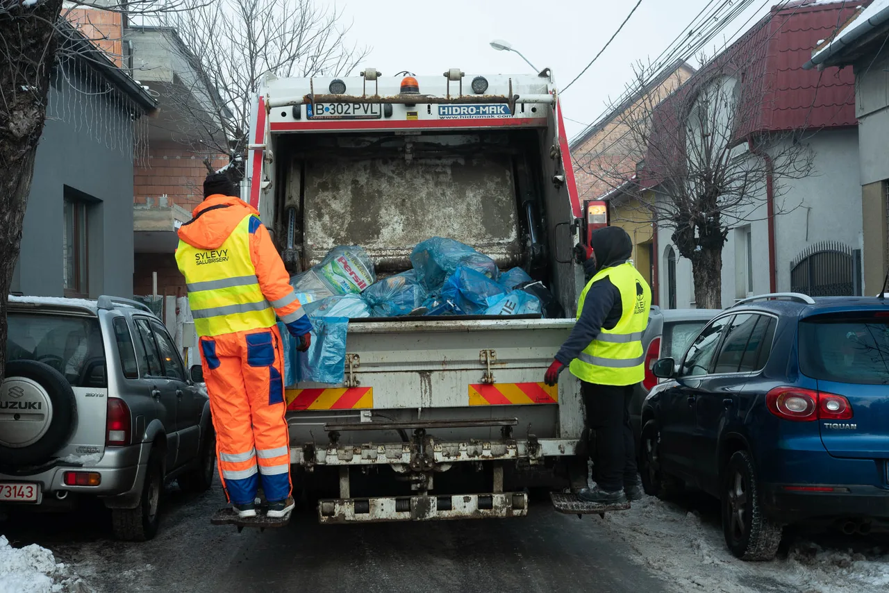 Két narancssárga és sárga láthatósági ruhát viselő szemétszállító dolgozó egy szemetes tehergépkocsi hátulján áll, kék szemeteszsákokat dobnak a felnyitott konténerbe egy havas, autókkal parkolt utcán.