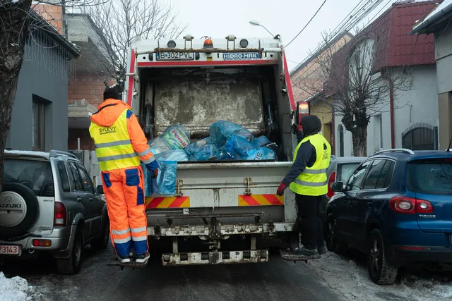 Két narancssárga és sárga láthatósági ruhát viselő szemétszállító dolgozó egy szemetes tehergépkocsi hátulján áll, kék szemeteszsákokat dobnak a felnyitott konténerbe egy havas, autókkal parkolt utcán.