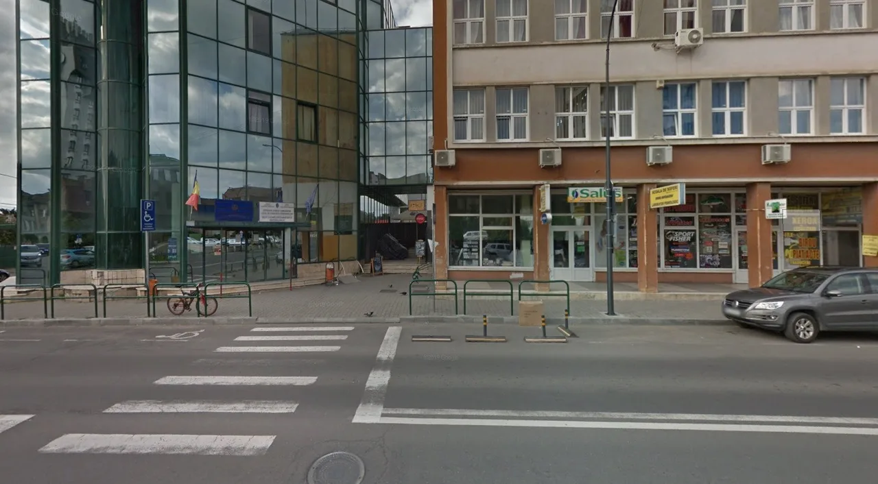 Utcasarok üvegfalú irodaépülettel és egy régebbi, boltokkal teli épülettel, előtte gyalogátkelő, parkoló autók és kerékpártárolók.