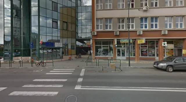 Utcasarok üvegfalú irodaépülettel és egy régebbi, boltokkal teli épülettel, előtte gyalogátkelő, parkoló autók és kerékpártárolók.