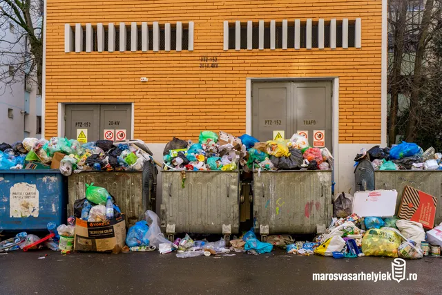Túltelt szemétkonténerek sorakoznak egy narancssárga téglaépület előtt, a konténerek mellett és tetején szétszóródva műanyag zsákok, kartondobozok és egyéb hulladék halmozódik fel.