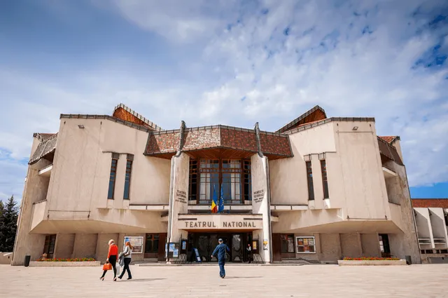 A Marosvásárhelyi Nemzeti Színház (Teatrul Național) épületének homlokzata látszik, előtte térrel, ahol néhány járókelő halad el.
