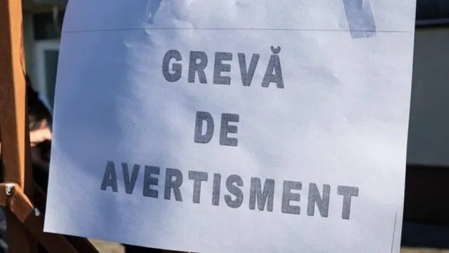Kézzel nyomtatott „GREVĂ DE AVERTISMENT” (figyelmeztető sztrájk) feliratú papírlap egy kerítésre vagy oszlopra ragasztva, kék ragasztószalaggal rögzítve.