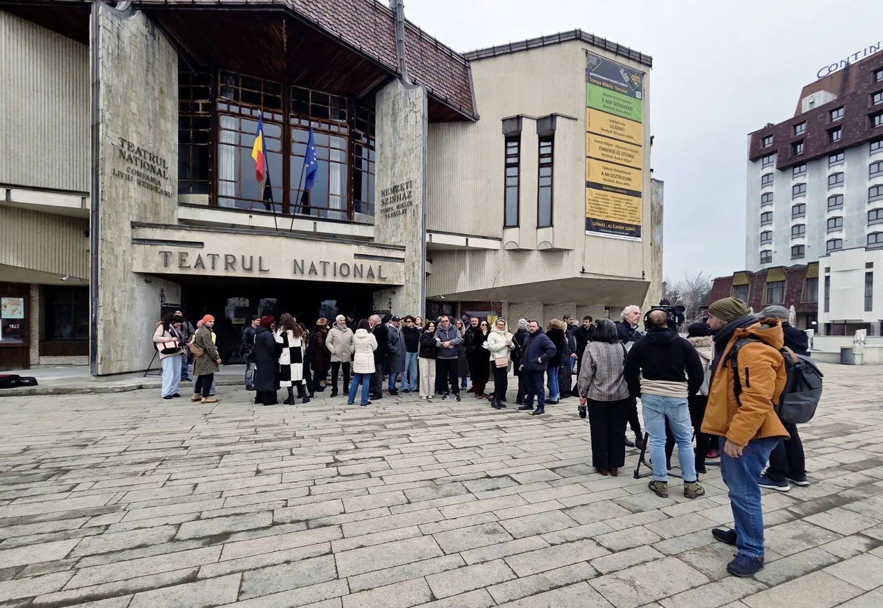 Emberek nagy csoportja gyűlik össze a marosvásárhelyi Nemzeti Színház bejárata előtt a téren, néhányan kamerával és mikrofonnal filmezik az összejövetelt.