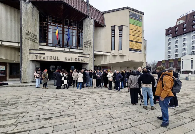 Emberek nagy csoportja gyűlik össze a marosvásárhelyi Nemzeti Színház bejárata előtt a téren, néhányan kamerával és mikrofonnal filmezik az összejövetelt.