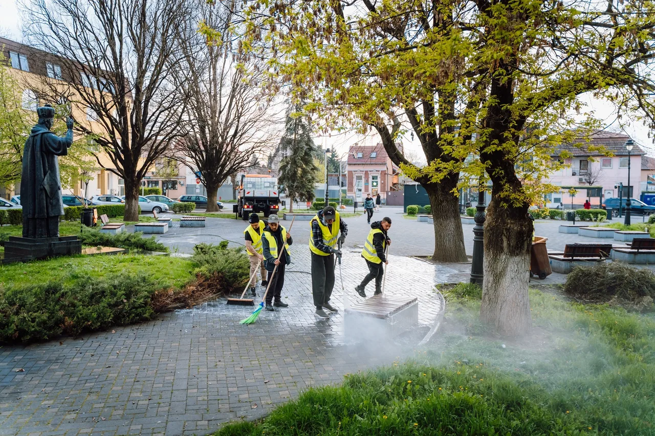Sárga láthatósági mellényt viselő köztisztasági dolgozók söprik és tisztítják egy park köves sétányát, a háttérben egy szobor és városi épületek láthatók.