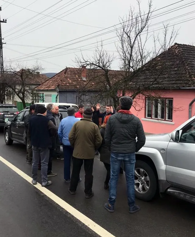 Kis csoport ember áll az utcán, autók között, falusi házsor előtt, láthatóan beszélgetnek vagy tájékozódnak egy helyi ügyről.