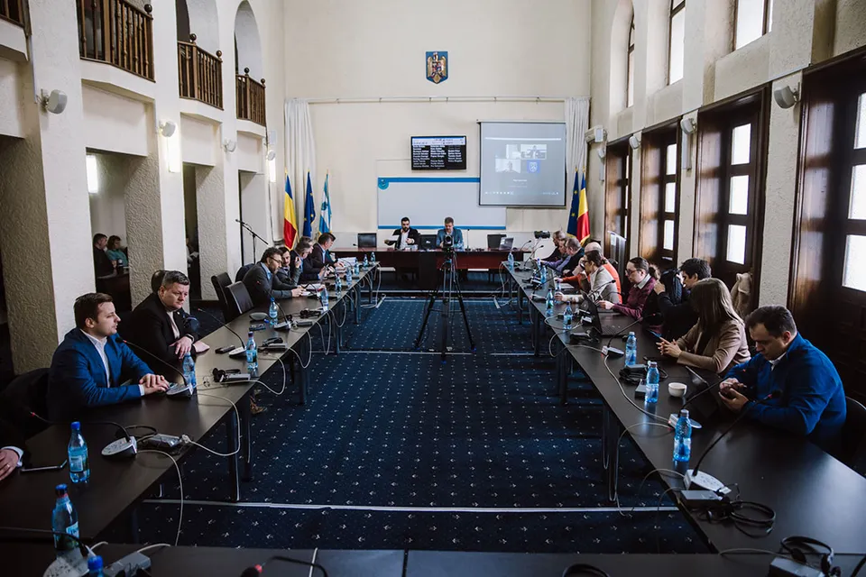 Hivatalos teremben ülés zajlik: két hosszú asztal mentén ülnek a jelenlevők, az asztalokon mikrofonok, vizes palackok és fülhallgatók, a hátsó falon vetítőképernyő, kijelző és zászlók, a falon pedig egy városi címer látható.