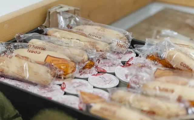 Fóliába csomagolt kukoricakenyerek (Corn Simplu) és kerek vajas vagy sajtos adagok egy dobozban, tálcán elrendezve.