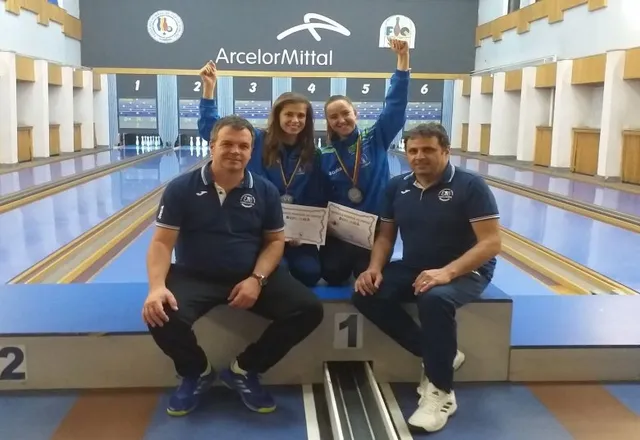 Két érmes fiatal sportolónő és két edző pózol egy dobogón egy tekepálya előtt, oklevelekkel a kezükben, kezüket ünnepelve a magasba emelve. A háttérben tekepályák és ArcelorMittal felirat látható.