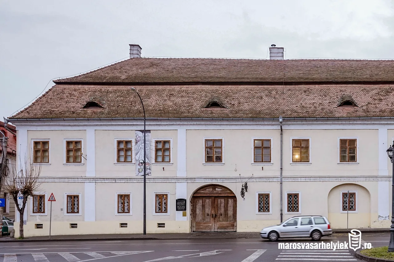 A Teleki-Bolyai Könyvtár (Teleki Téka) épületének utcai homlokzata látható, cserepes magastetővel, három kis tetőablakkal, fehér falú front homlokzaton sárgás-fehér színezéssel, íves fa kapubejárattal és több ablakkal; előtte az utca és egy elhaladó ezüstszínű autó.