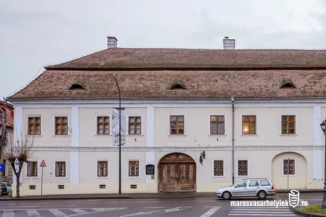 A Teleki-Bolyai Könyvtár (Teleki Téka) épületének utcai homlokzata látható, cserepes magastetővel, három kis tetőablakkal, fehér falú front homlokzaton sárgás-fehér színezéssel, íves fa kapubejárattal és több ablakkal; előtte az utca és egy elhaladó ezüstszínű autó.