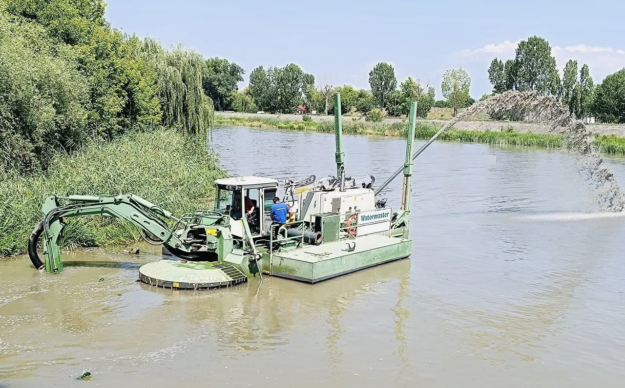 Egy Watermaster típusú úszó munkagép dolgozik egy folyó medrében, kotrókaros elülső szerkezettel a víz alatti iszap eltávolítására, miközben a felszívott anyagot a partra fröccsenti. A gépben egy kezelő ül, mellette egy kék ruhás dolgozó áll.
