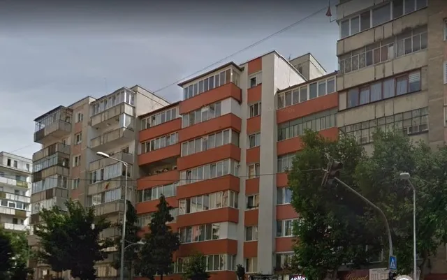 Panelházak sora látható egy városi utcában, előttük fák és utcai lámpák, jellemzően erkélyes homlokzatokkal, piros-fehér csíkos elemekkel.