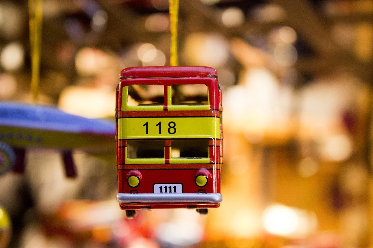 Egy vörös-sárga színű, fém miniatura autóbusz lóg egy vékony sárga zsinóron, valószínűleg ékszerként vagy fülbevalóként. A busz oldalán a 118-as szám, elején pedig egy fehér táblán a 1111-es szám olvasható; a háttér erősen elmosódott, meleg, sárgás tónusú.