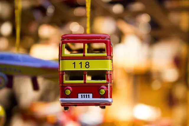 Egy vörös-sárga színű, fém miniatura autóbusz lóg egy vékony sárga zsinóron, valószínűleg ékszerként vagy fülbevalóként. A busz oldalán a 118-as szám, elején pedig egy fehér táblán a 1111-es szám olvasható; a háttér erősen elmosódott, meleg, sárgás tónusú.