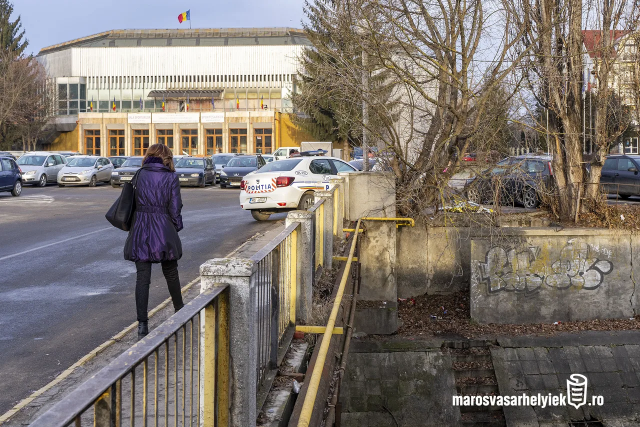 Utcakép a Sportcsarnok előtti parkolóval, ahol egy nő lila kabátban sétál egy korlátos híd mellett, előtte rendőrségi autó parkol. A híd alatt egy csatorna vagy vízfolyás medre látszik, firkált betonfallal.