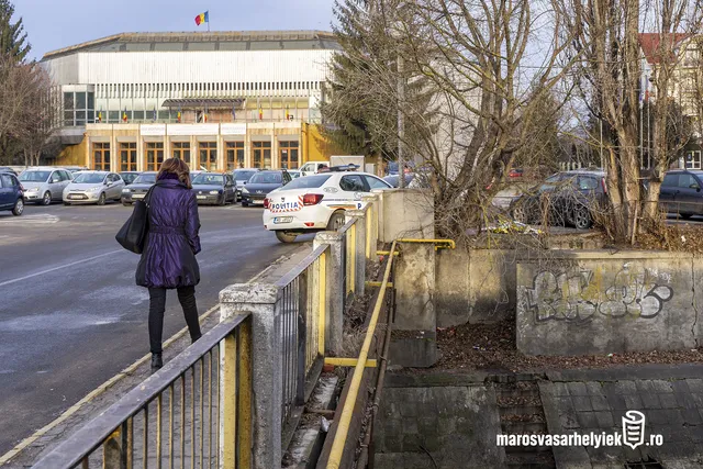Utcakép a Sportcsarnok előtti parkolóval, ahol egy nő lila kabátban sétál egy korlátos híd mellett, előtte rendőrségi autó parkol. A híd alatt egy csatorna vagy vízfolyás medre látszik, firkált betonfallal.